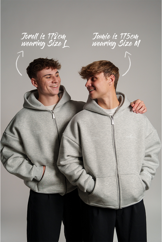 Zwei Männer tragen einen hellgrauen Zip-Hoodie. Links steht Jorell (178 cm, Größe L), rechts Jamie (175 cm, Größe M). Beide lächeln sich an und zeigen den lockeren Sitz des Hoodies mit Kapuze und Reißverschluss.