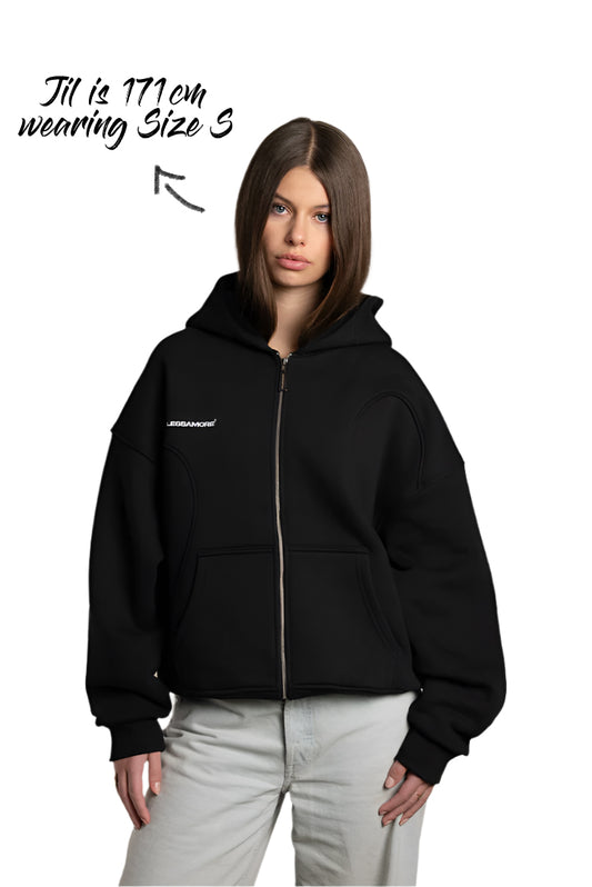 Jil trägt einen schwarzen Zip-Hoodie mit weißem Brustprint „ENDLESSAMORE®“. Die Kapuze ist locker aufgesetzt, der Reißverschluss ist geschlossen. Der Hoodie ist cropped geschnitten und hat sichtbare Teilungsnähte.