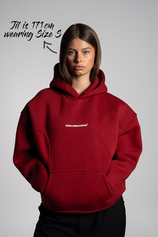 Frau mit geradem, braunem Haar trägt einen bordeauxroten Hoodie mit weißem „ENDLESSAMORE“-Logo auf der Brust. Der Hoodie hat eine strukturierte Schnittführung im Schulterbereich.