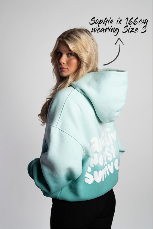 Rückansicht eines mintgrünen Oversized-Hoodies mit Farbverlauf, getragen von einer Frau. Auf dem Rücken ist ein großer, weißer 3D-Puffprint mit dem Statement „wanna live not just survive“ in verspielter Typografie zu sehen.