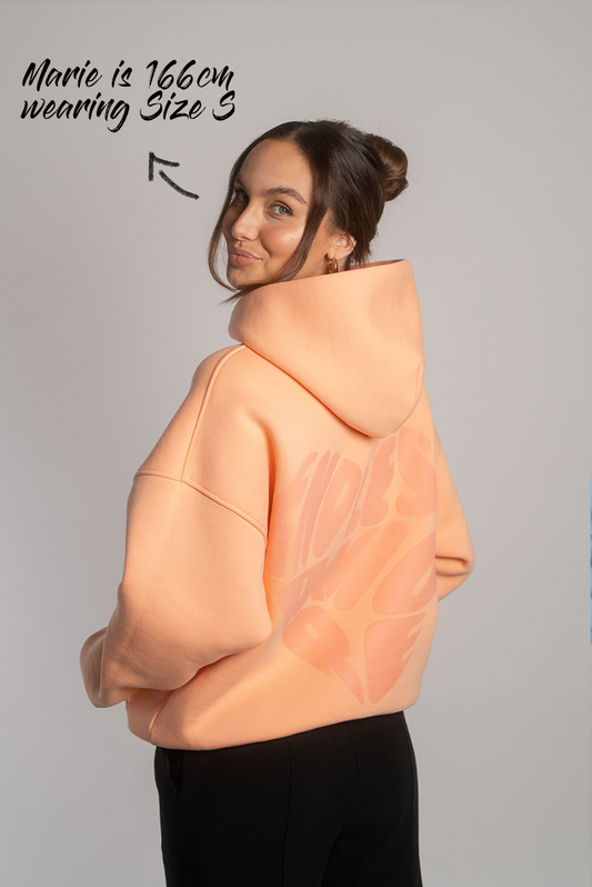 Apricotfarbener Hoodie mit tonalem Herz-Rückendruck „ENDLESS AMORE“, getragen von Frau mit hochgesteckten Haaren, seitlich von hinten gezeigt.