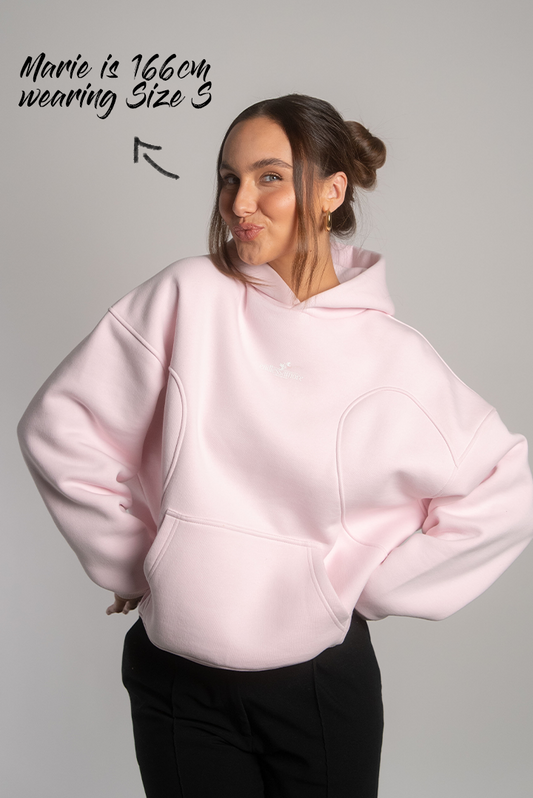 Rosa Kapuzenpullover mit formgebenden Teilungsnähten auf Brusthöhe, Kängurutasche und kleinem „endlessamore“-Logo in Weiß auf der Brust, getragen von einem Model mit schwarzer Hose.