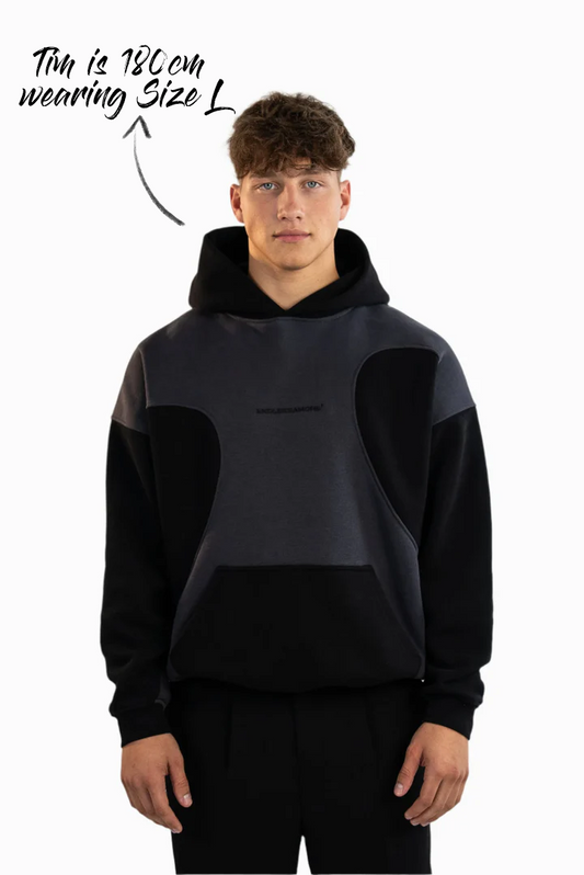 Schwarz-grauer Kapuzenpullover mit kontrastierenden Stoffeinsätzen und Kängurutasche, getragen von einem Model mit dunkler Hose, stehend in Frontalansicht.