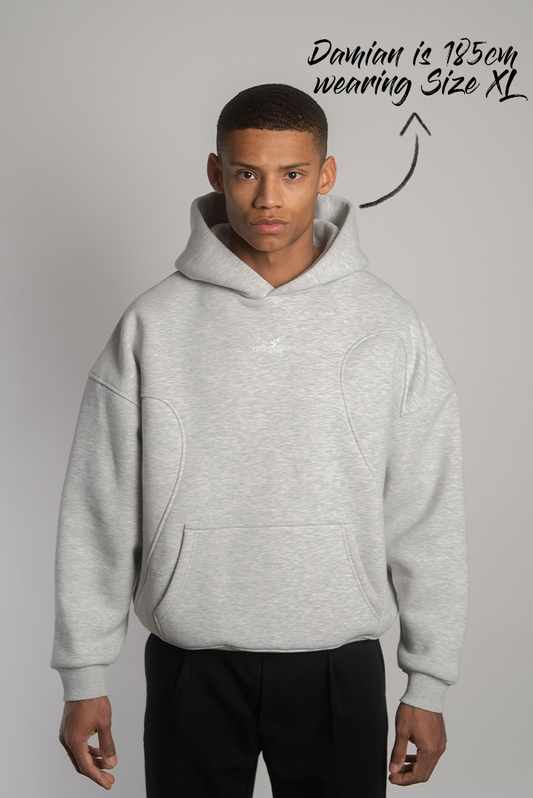 Grauer Kapuzenpullover mit dezentem weißen „endlessamore“-Frontprint in Brusthöhe, getragen von einer Person mit leicht ernster Mimik. Sichtbar sind überschnittene Schultern, markante Teilungsnähte und eine Kängurutasche.