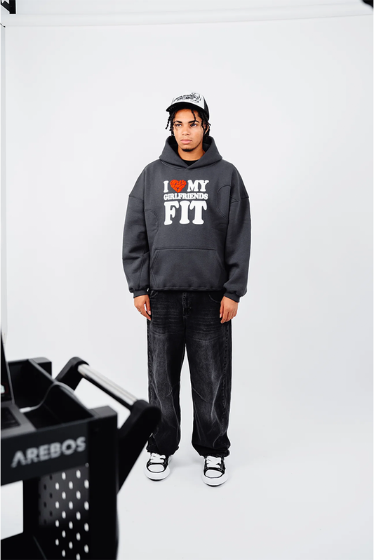 Person steht frontal in einem locker sitzenden, anthrazitfarbenen Hoodie mit dem Aufdruck „I ♥ MY GIRLFRIENDS FIT“ in Weiß und Rot. Dazu werden verwaschene, schwarze Baggy-Jeans und schwarz-weiße Sneaker getragen. Im Vordergrund links ist verschwommen ein schwarzer Studio-Wagen zu sehen. Der Hintergrund ist weiß.