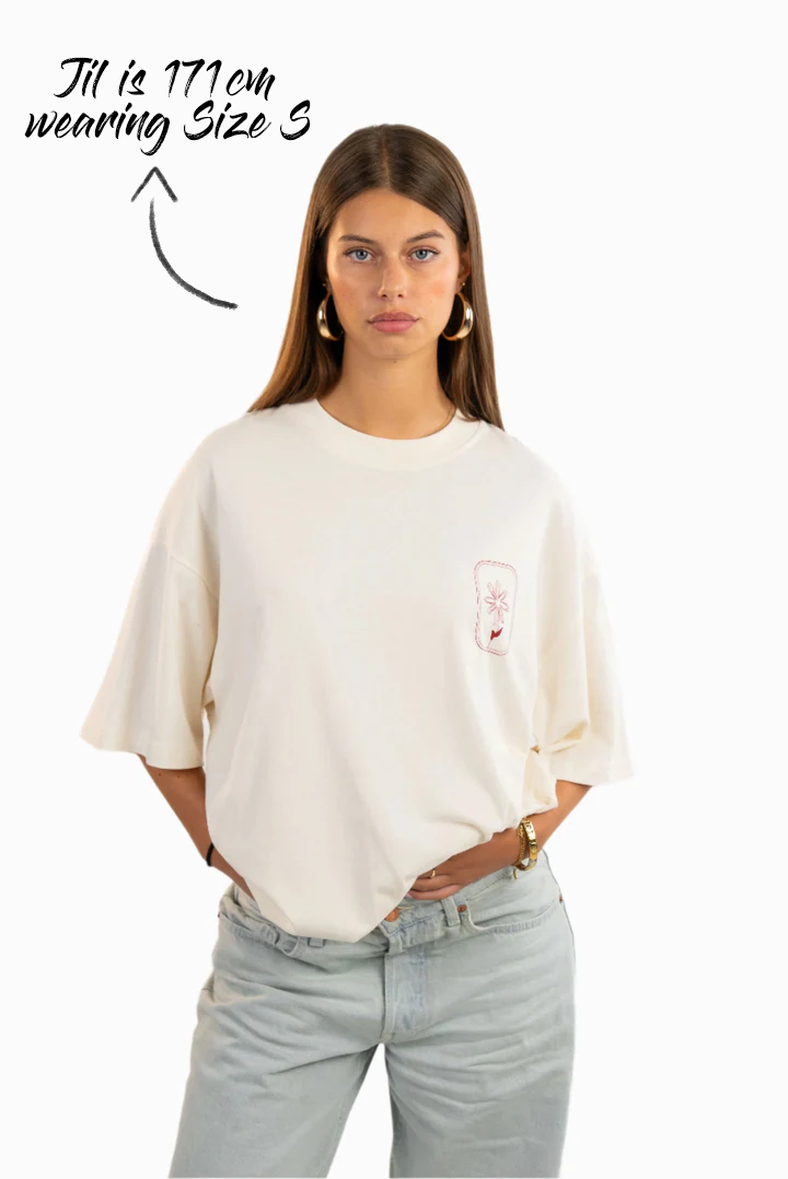 Frontalansicht eines weißen T-Shirts mit kleiner roter Blumenstickerei auf der linken Brust. Getragen von einer Frau in Größe S (171 cm groß).
