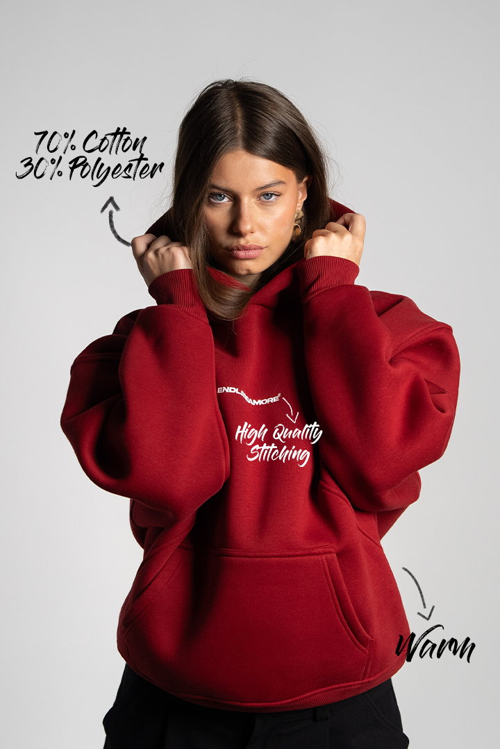 Frau trägt den bordeauxroten Hoodie mit beiden Händen am Kapuzenkragen. Zu sehen sind der weiße Brustprint und die Materialstruktur. Textgrafiken deuten auf Material und Qualität hin.