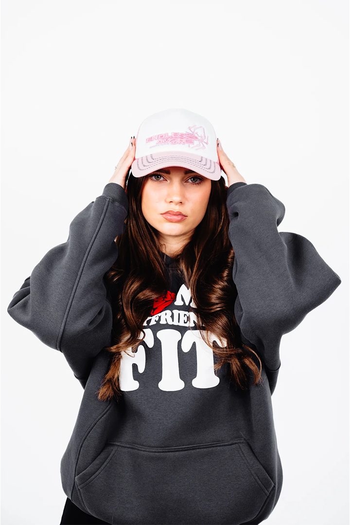 Frau blickt direkt in die Kamera, beide Hände fassen an die pink-weiße Trucker-Cap mit gesticktem „ENDLESS AMORE“-Schriftzug. Der Hoodie mit weißem Frontprint ist locker geschnitten.