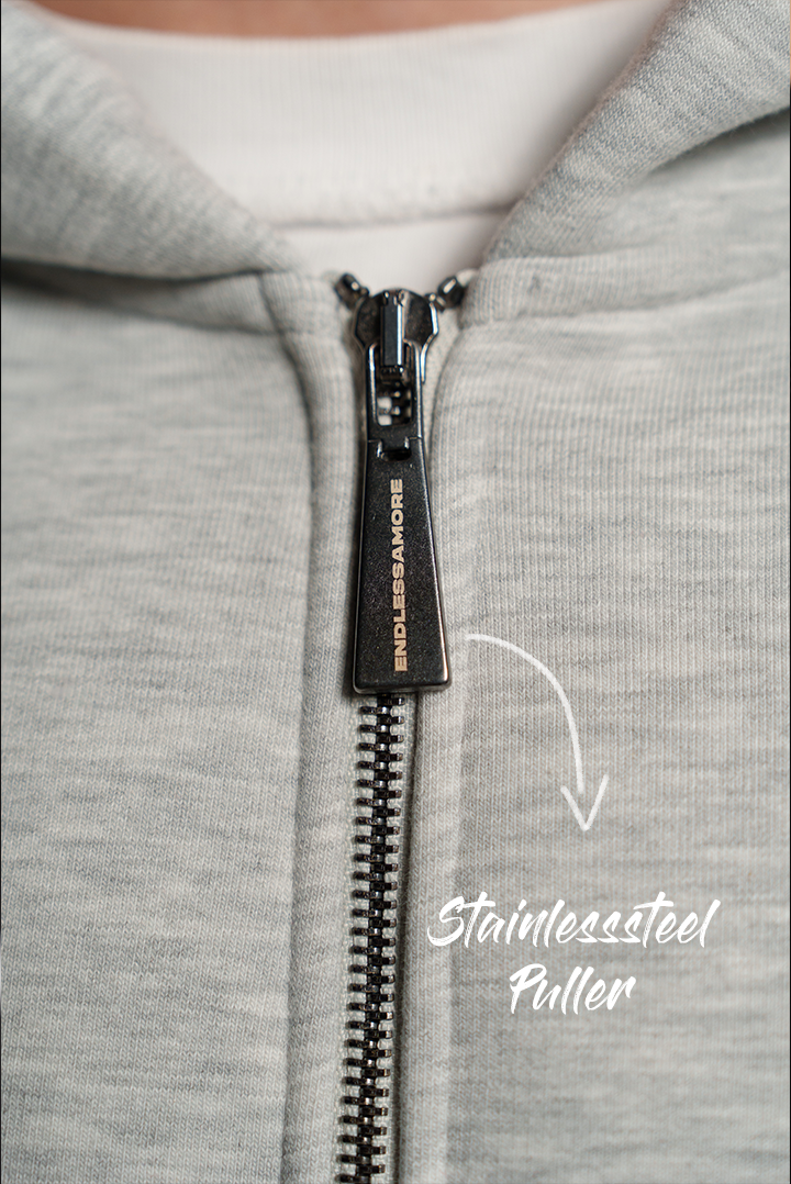 Detailansicht des Reißverschlusses mit graviertem Zipper aus Metall. Auf dem Zipper steht „ENDLESSAMORE“. Der Reißverschluss verläuft mittig entlang des grauen Hoodies.