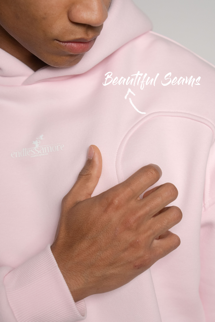 Nahaufnahme der Brustpartie eines rosa Hoodies mit weißen Nähten in Herzform und dezentem Logo, Finger berührt die Naht leicht.