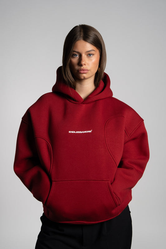 Frau mit geradem, braunem Haar trägt einen bordeauxroten Hoodie mit weißem „ENDLESSAMORE“-Logo auf der Brust. Der Hoodie hat eine strukturierte Schnittführung im Schulterbereich.