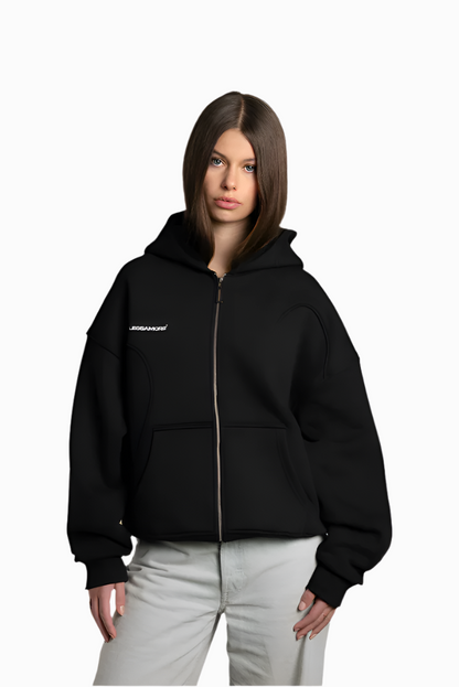 Jil trägt einen schwarzen Zip-Hoodie mit weißem Brustprint „ENDLESSAMORE®“. Die Kapuze ist locker aufgesetzt, der Reißverschluss ist geschlossen. Der Hoodie ist cropped geschnitten und hat sichtbare Teilungsnähte.