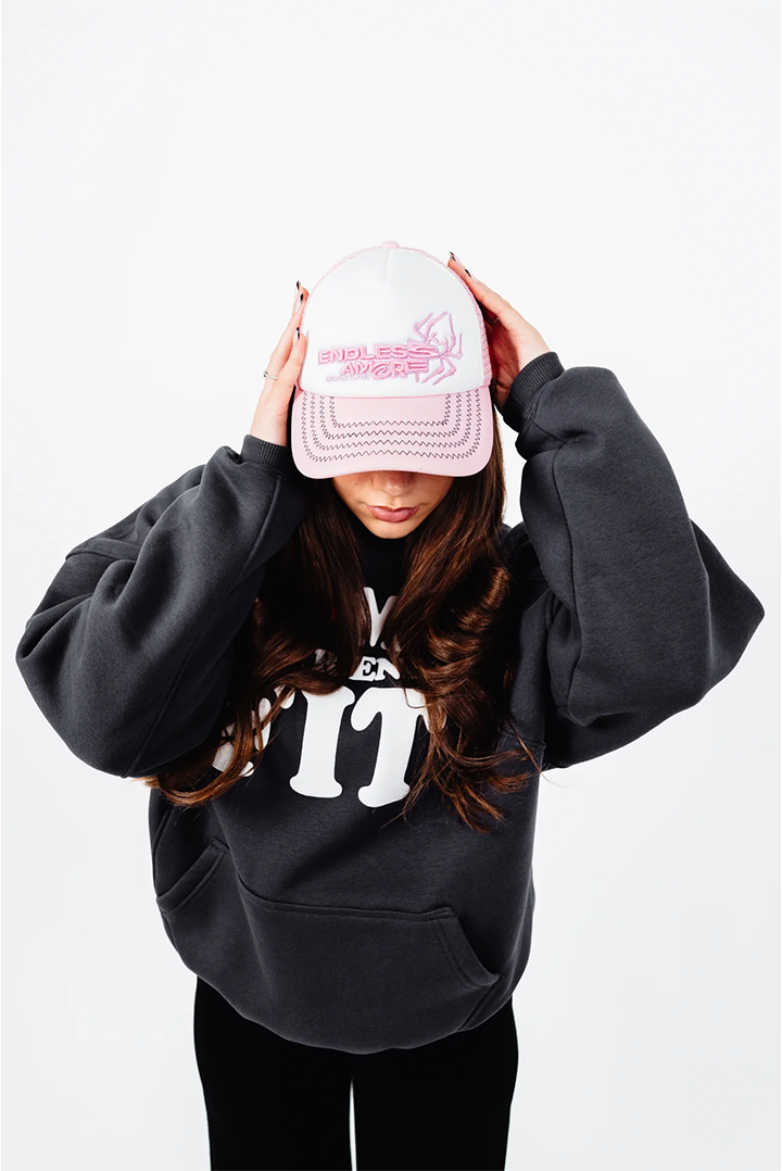 Nahaufnahme einer Person mit langen, gewellten Haaren, die eine pink-weiße Trucker-Cap mit dem gestickten Schriftzug „ENDLESS AMORE“ und einem grafischen Herzmotiv trägt. Die Cap hat einen weißen Frontbereich und pinke Nähdetails am Schirm. Die Person trägt einen dunkelgrauen Hoodie mit weißem Druck.