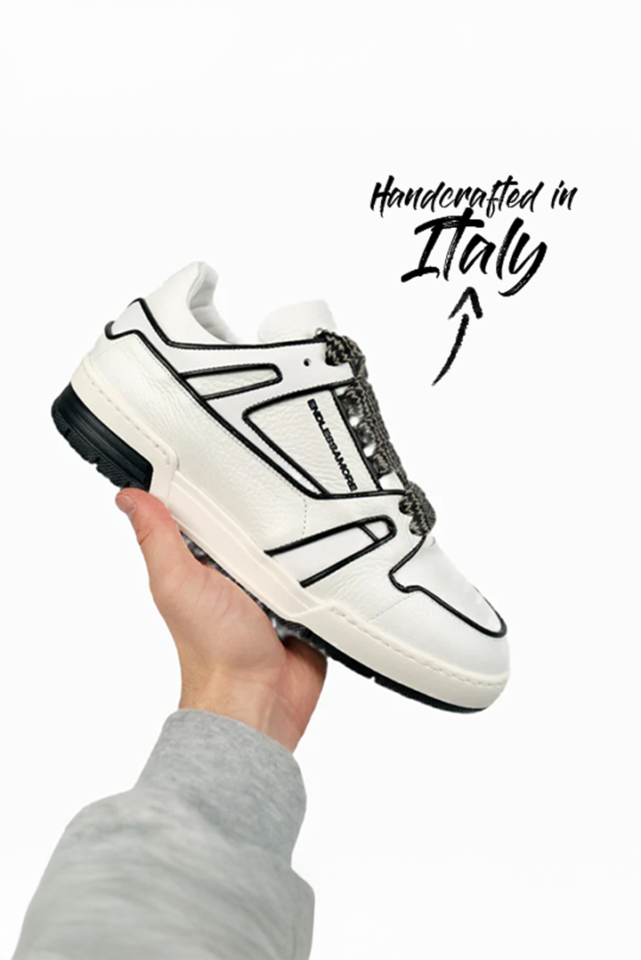 Weißer Sneaker mit schwarzer Umrandung und strukturiertem Leder, von einer Hand vor hellem Hintergrund präsentiert. Die schwarze Sohle und gemusterte Schnürsenkel ergänzen das Design.