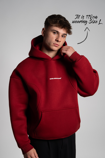 Mann mit kurzem Haar trägt einen bordeauxroten Hoodie mit zentral platziertem weißen „ENDLESSAMORE“-Brustprint. Der Hoodie hat betonte Nahtführung im Brustbereich.