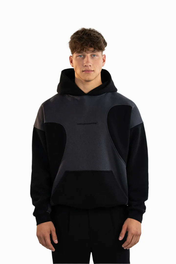 Schwarz-grauer Kapuzenpullover mit kontrastierenden Stoffeinsätzen und Kängurutasche, getragen von einem Model mit dunkler Hose, stehend in Frontalansicht.