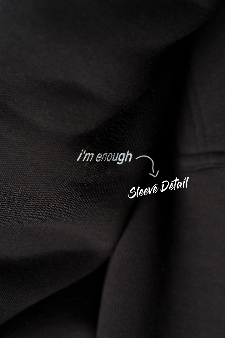  Nahaufnahme des schwarzen Ärmels mit weißem Textdruck „i’m enough“. Daneben ist die Beschriftung „Sleeve Detail“ als handschriftliche Grafik ergänzt.