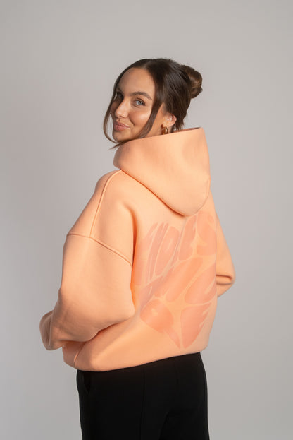 Apricotfarbener Hoodie mit tonalem Herz-Rückendruck „ENDLESS AMORE“, getragen von Frau mit hochgesteckten Haaren, seitlich von hinten gezeigt.