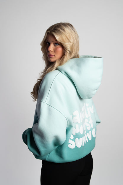 Rückansicht eines mintgrünen Oversized-Hoodies mit Farbverlauf, getragen von einer Frau. Auf dem Rücken ist ein großer, weißer 3D-Puffprint mit dem Statement „wanna live not just survive“ in verspielter Typografie zu sehen.