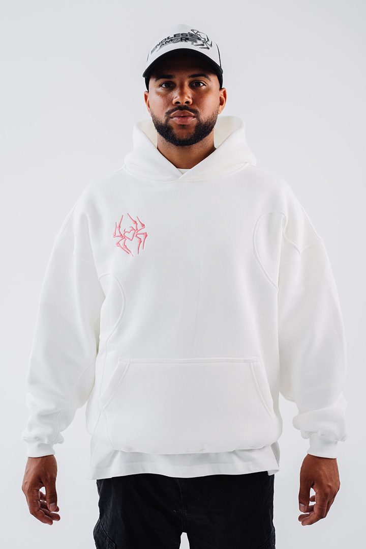Weißer Hoodie mit rosaner Stickerei in Form eines stilisierten Herzens auf der linken Brustseite. Der Hoodie hat sichtbare Teilungsnähte und eine Kängurutasche. Getragen wird er locker über einer weiteren Lage, kombiniert mit einer schwarz-weißen Cap und schwarzer Hose. Der Hintergrund ist hell und neutral gehalten.